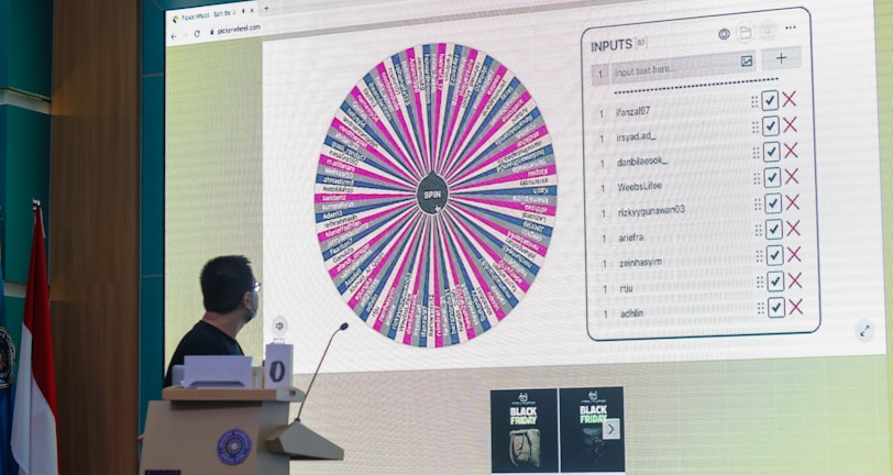 A person stands in front of a large screen displaying a colorful digital wheel with names. The screen shows a web interface with options for inputs, featuring a list of names with checkboxes on one side and a large, vibrant spinning wheel on the other. The environment appears to be indoors, possibly in a conference or presentation setting with a podium.